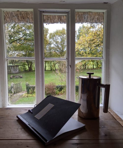 A french press coffee pot and journal sit on a desk looking out into the garden